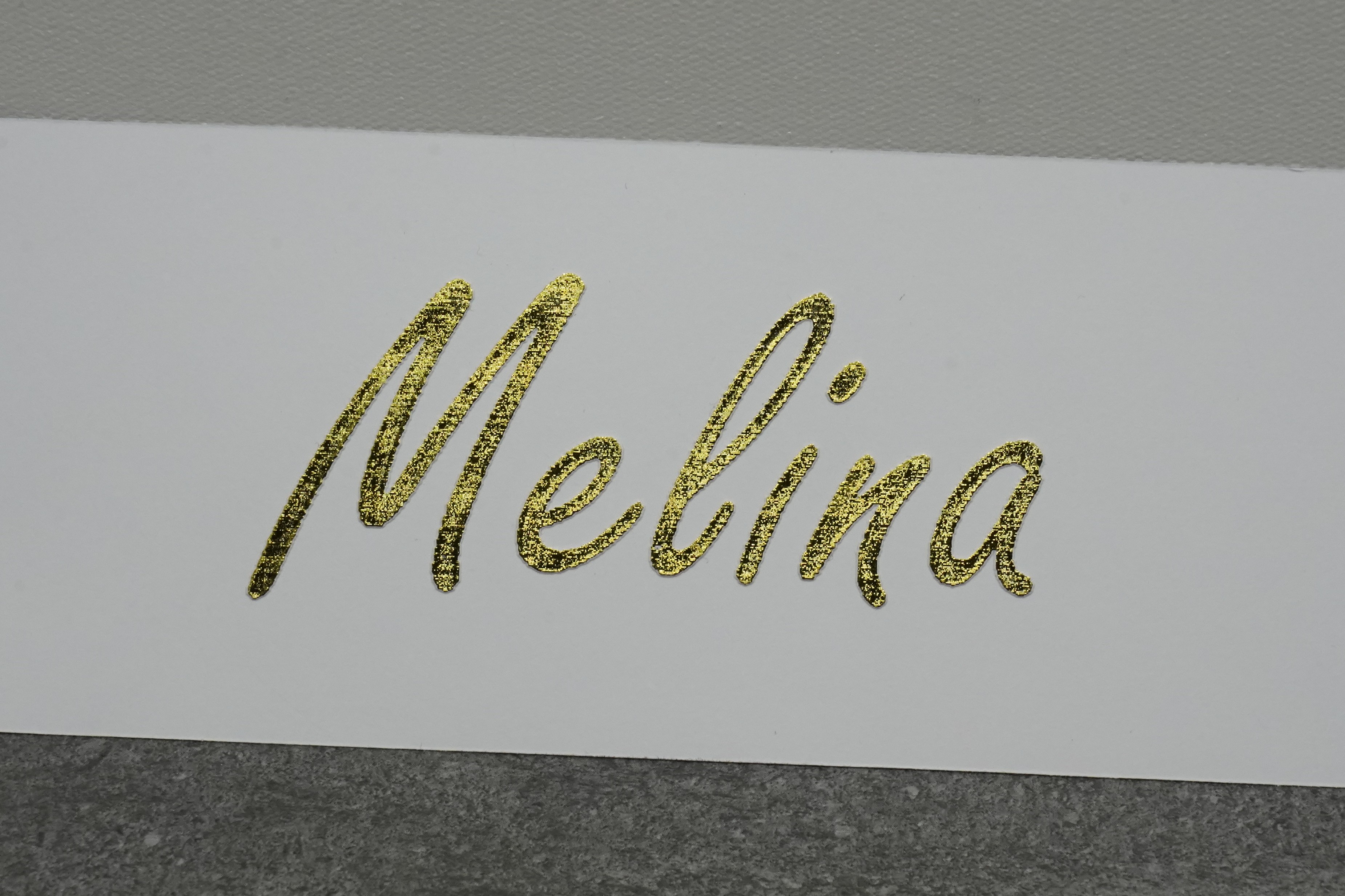 Makro-Aufnahme des Vornamens «Melina» in echter Goldfolie auf weissem Papier — glitzernde, körnig-metallische Oberfläche, feine Kalligraphie-Kurven in Handschrift-Optik